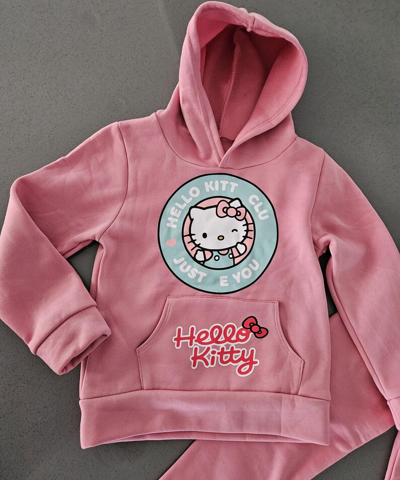 K&ouml;nnte beinhalten: Rosa Hello Kitty Hoodie und passende Hose. Der Hoodie hat eine Vordertasche und eine runde Grafik mit Hello Kitty und dem Text "Hello Kitt Club Just E You". Das Hello Kitty-Logo befindet sich ebenfalls auf der Vordertasche.