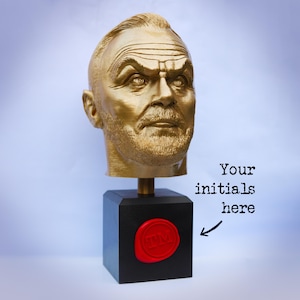 May include: A gold-colored bust of a man's head atop a black square base. The base features a red wax seal with initials and the text "Your initials here". The bust is detailed with facial features and hair.