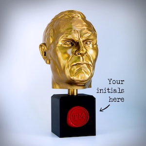 May include: A gold-colored sculpture of a human head mounted on a black square base. The base features a circular red wax-style seal with the letters TM embossed in the center. Text to the right reads: Your initials here with an arrow.