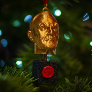 May include: A gold-colored Christmas ornament shaped like a bust of a man's head. The ornament is attached to a black cube with a red wax seal. The ornament is hanging from a red ribbon against a blurred background of Christmas tree lights.