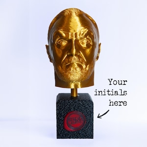 Może przedstawiać: Złoty popiersie głowy mężczyzny na czarnej, teksturowanej podstawie. Podstawa posiada czerwoną pieczęć z inicjałami "TM" i tekstem "Your initials here". Popiersie to szczegółowa rzeźba z brodą i zmarszczonymi brwiami.