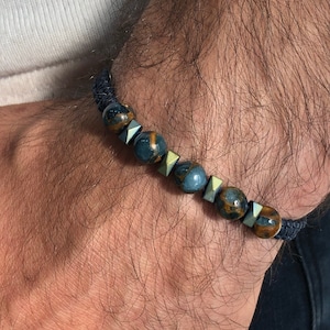 Può includere: Un braccialetto con perline blu e dorate con un cordoncino nero. Le perline sono rotonde e hanno un motivo maculato.
