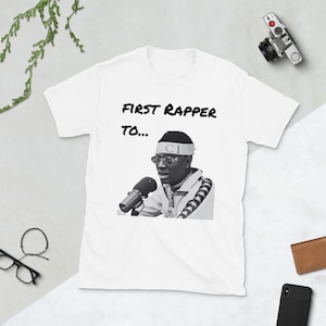 May include: White t-shirt with a black and white image of a rapper wearing a Gucci headband and a microphone. The text "FIRST RAPPER TO..." is printed above the image.