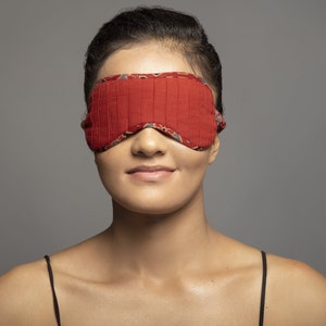 以下が含まれることがあります： プリーツデザインの赤いスリープマスクが目を覆っています。マスクの縁には、グレー、赤、白のディテールが施された模様の生地が使用されています。背景は無地のグレーです。