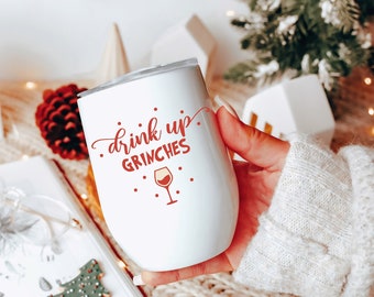 以下が含まれることがあります： 「drink up, Grinches」と赤文字で書かれた白いワインタンブラー。タンブラーには、赤ワインの入った赤ワイングラスのアイコンがあります。