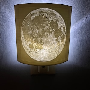 May include: A white night light shaped like a crescent moon with a detailed image of the moon's surface. The night light is plugged into an electrical outlet.