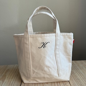 Puede incluir: Bolso tote de lona color crema con dos asas y una letra "K" bordada en negro. La bolsa está sobre una superficie de madera. El bolso tote es de color neutro.