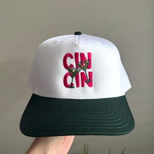 Pode incluir: Boné de beisebol branco e verde escuro com a palavra "CIN CIN" bordada em rosa, juntamente com dois copos de martini. O boné tem uma aba plana e uma parte inferior verde escuro.