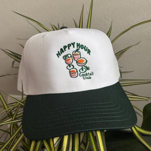 Peut inclure: Casquette de baseball blanche et vert foncé avec les mots "HAPPY HOUR Cocktail Club" brodés en vert. La casquette présente une illustration de trois cocktails. La visière est vert foncé.