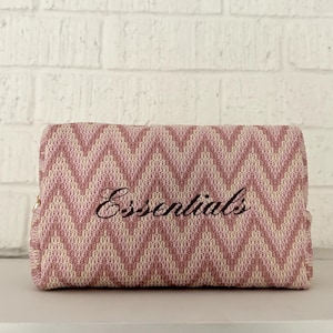 May include: A rectangular, pink and white chevron-patterned cosmetic bag. The word "Essentials" is embroidered in a cursive font across the front. The bag is closed and appears to be made of a woven fabric.