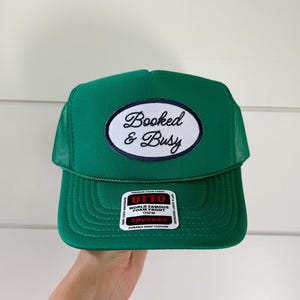 May include: Green trucker hat with the text "Booked & Busy" on a white oval patch. The hat has a mesh back and a foam front. The tag reads "World Famous Foam Front".
