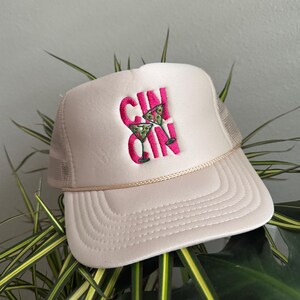 Pode incluir: Boné de caminhoneiro rosa claro com parte traseira em tela e aba curva. O boné apresenta o texto bordado "GIN" em rosa brilhante, com um design de copo de coquetel. Um detalhe de corda bege adorna a aba.