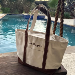 Puede incluir: Bolso tote de lona color crema con detalles marrones, incluyendo la base, las asas y los ribetes. El bolso tiene un bolsillo frontal y el texto bordado "on the go!". El bolso está sobre una superficie de madera cerca de una piscina.