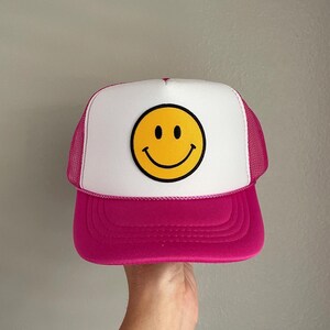 May include: A pink and white trucker hat with a yellow smiley face patch. The hat has a mesh back.