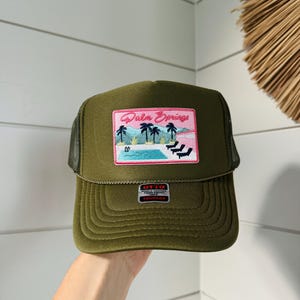 Könnte beinhalten: Olivgrüne Trucker-Mütze mit Mesh-Rückseite und strukturierter Vorderseite. Die Mütze hat einen rechteckigen Aufnäher mit den Worten "Palm Springs" über einer Poolszene mit Palmen und Liegestühlen. Die Mütze hat einen gebogenen Schirm.