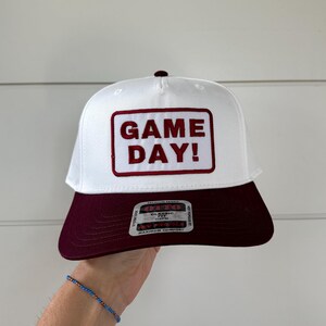 Może przedstawiać: Biała czapka bejsbolowa z bordowym daszkiem i bordowym kwadratem z napisem "GAME DAY!" bordowymi literami. Czapka jest trzymana przez osobę.
