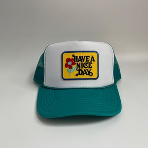 Puede incluir: Gorra de camionero blanca y turquesa con un panel frontal blanco y una visera turquesa. El sombrero presenta un parche con las palabras "HAVE A NICE DAY" y un diseño floral. La parte trasera de malla es turquesa.