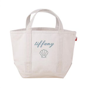 May include: Off-white canvas tote bag with two handles. The word "tiffany" is embroidered in teal script above a teal seashell design. A small red tag is attached to the side of the bag.