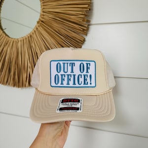 Puede incluir: Una gorra de camionero beige con un parche azul que dice "OUT OF OFFICE!" en letras blancas. La gorra tiene una etiqueta negra "OTTO" con el texto "WORLD FAMOUS FOAM FRONT" y "TRUCKER" en ella.
