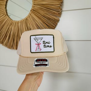 May include: A beige trucker hat with a white patch displaying a martini glass and the words "tini time." The hat has a mesh back and a curved peak. A small tag is visible on the brim.