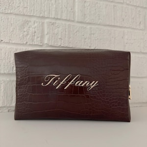 Puede incluir: Un neceser rectangular marrón oscuro con textura de piel de cocodrilo. El nombre "Tiffany" está bordado en elegante escritura cursiva en la parte delantera. La bolsa tiene un cierre de cremallera dorado.