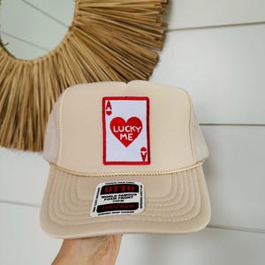 May include: A beige trucker hat with a red and white patch that says "Lucky Me" and features a red heart and an ace of hearts. The hat has a black and white label that says "Otto World Famous Foam Front OSFM Trucker Durable Snap Closure."