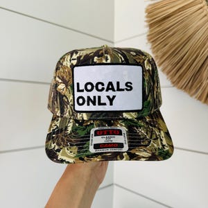 May include: A camouflage trucker hat with a white patch that says "Locals Only" in black text. The hat has a red label that says "Otto" in white text and "Classic Fit One Size" in black text.