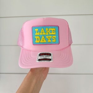 May include: Pink trucker hat with a blue and yellow patch that says "LAKE DAYS". The hat has a black label that says "OTTO WORLD FAMOUS FOAM FRONT OSFM TRUCKER DURABLE SNAP CLOSURE".