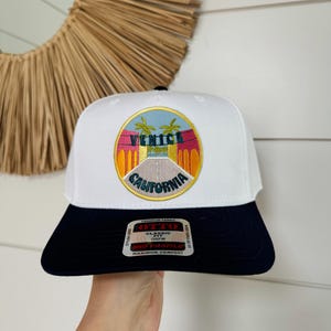 Op de afbeelding: Witte en marineblauwe baseballpet met een kleurrijk geborduurd patch met de tekst "Venice California". De patch toont palmbomen en een weg. De pet heeft een klassieke pasvorm en een middelhoog profiel.