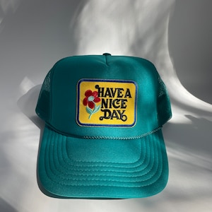 May include: A teal blue trucker hat with a yellow patch that says "HAVE A NICE DAY" with a red and blue flower graphic.