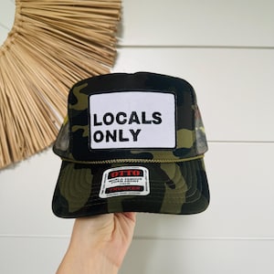 May include: A camouflage trucker hat with a white patch that reads "LOCALS ONLY" in black block letters. The hat has a mesh back and a curved brim. The hat is a snap closure.