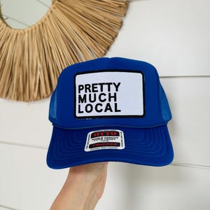 May include: A royal blue trucker hat with a white patch that says "Pretty Much Local" in black text. The hat has a black mesh back and a red Otto trucker label.