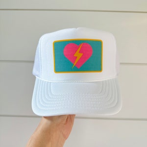 Peut inclure: Casquette de camionneur blanche avec un patch carré bleu turquoise représentant un cœur rose avec un éclair jaune à l'intérieur.