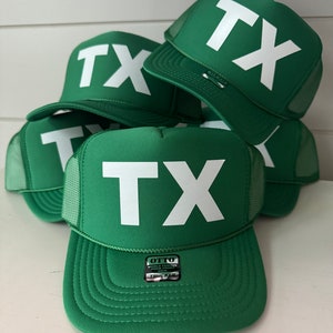 May include: A stack of green trucker hats with a white "TX" logo on the front. The hats have a mesh back and a black label that says "OTTO WORLD FAMOUS TRUCKER CAP DFM TRUCKER".