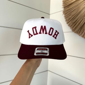 Könnte beinhalten: Weiß-maroonfarbene Baseballkappe mit dem Wort "HOWDY" in maroonfarbenen Blockbuchstaben. Die Kappe hat einen flachen Schirm und eine klassische Passform. Die Kappe ist aus hochwertigem Stoff für maximalen Komfort gefertigt.