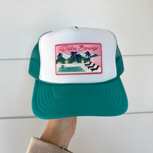 May include: A white and green trucker hat with a pink and green embroidered patch that says "Palm Springs" and depicts palm trees and a pool.