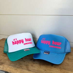 May include: Two trucker hats with the text "It's happy hour somewhere!" in pink lettering. One hat is green and white, the other is blue and white.