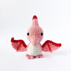 May include: A pink and red crocheted pterodactyl toy with black eyes. The toy has a white belly and red wings.