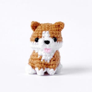 May include: A small crocheted stuffed dog with brown and white fur. The dog has black eyes and a pink tongue.