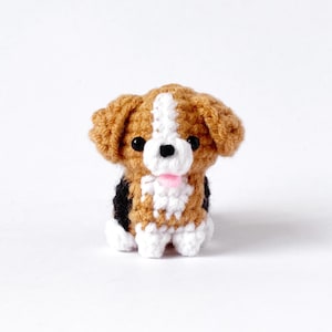 May include: A crocheted dog ornament in brown, white, and black. The dog has a pink tongue and black eyes. The dog is sitting up, with its ears perked. The dog is made of yarn.