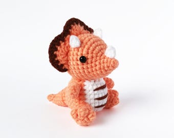 May include: A crocheted orange and brown triceratops dinosaur toy with white accents. The toy has black eyes and a small, white horn on its nose.