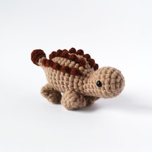 Op de afbeelding: Een gehaakt bruin en rood dinosaurus speelgoed met zwarte ogen.