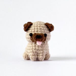 Pode incluir: Um brinquedo de cachorro pug de crochê com orelhas marrons, olhos pretos e uma língua rosa para fora. O brinquedo é feito de fio bege e está sentado em um fundo branco.