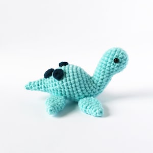 May include: A crocheted turquoise dinosaur toy with black eyes and spots. The dinosaur is lying on its belly with its head turned to the side.