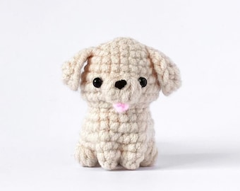 May include: A small, crocheted dog figurine in a sitting position. The dog is beige with black eyes and a pink tongue. The ears are floppy, and the body is made of textured yarn. The background is plain white.