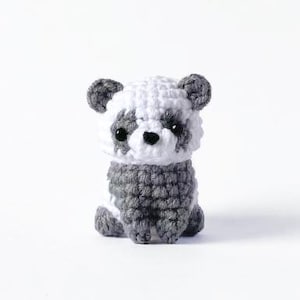 Pode incluir: Um urso panda de crochê feito à mão, em tons de branco e cinza. O panda tem olhos pretos pequenos e um nariz preto. O urso está sentado em posição vertical, mostrando seu trabalho de crochê detalhado. Um item charmoso e artesanal.