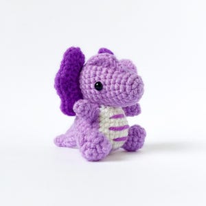 May include: A handmade crochet dragon in shades of purple, with a white belly featuring purple stripes. The dragon has large, dark eyes and a pair of purple wings. The toy is sitting on a white background.