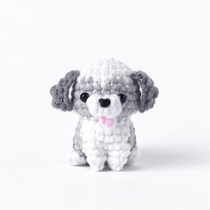 Puede incluir: Una figura de perro de ganchillo hecha a mano en blanco, gris y negro. El perro tiene ojos negros, orejas grises y una lengua rosa. El perro de ganchillo está sentado erguido y tiene un aspecto esponjoso.