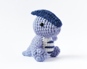 May include: A small, crocheted dinosaur toy with a blue hat and white stripes. The dinosaur is light purple and has black eyes.
