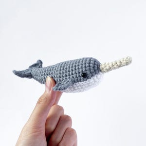May include: A gray and white crocheted narwhal plush toy with a small black eye. The toy is being held by a hand.